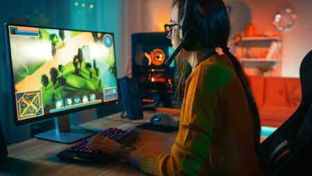 Excited and Concentrated Gamer Girl in Glasses and Headset with a Mic Playing Online Strategy Video Game on Her Personal Computer. Room and PC have Colorful Warm Neon Led Lights. Cozy Evening at Home.